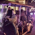 People riding an open-air tour cart decorated with purple lights, waving and smiling during a nighttime city event.